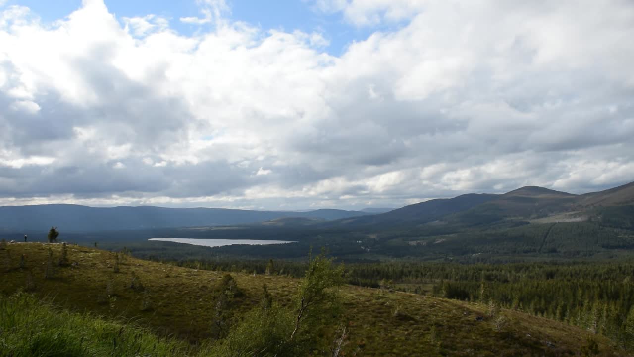 Wide view over green hill with scattered tree, large lake, mountain line, and bright cloud sky, showing peaceful outdoor scene with fresh air and natural beauty in open highland area