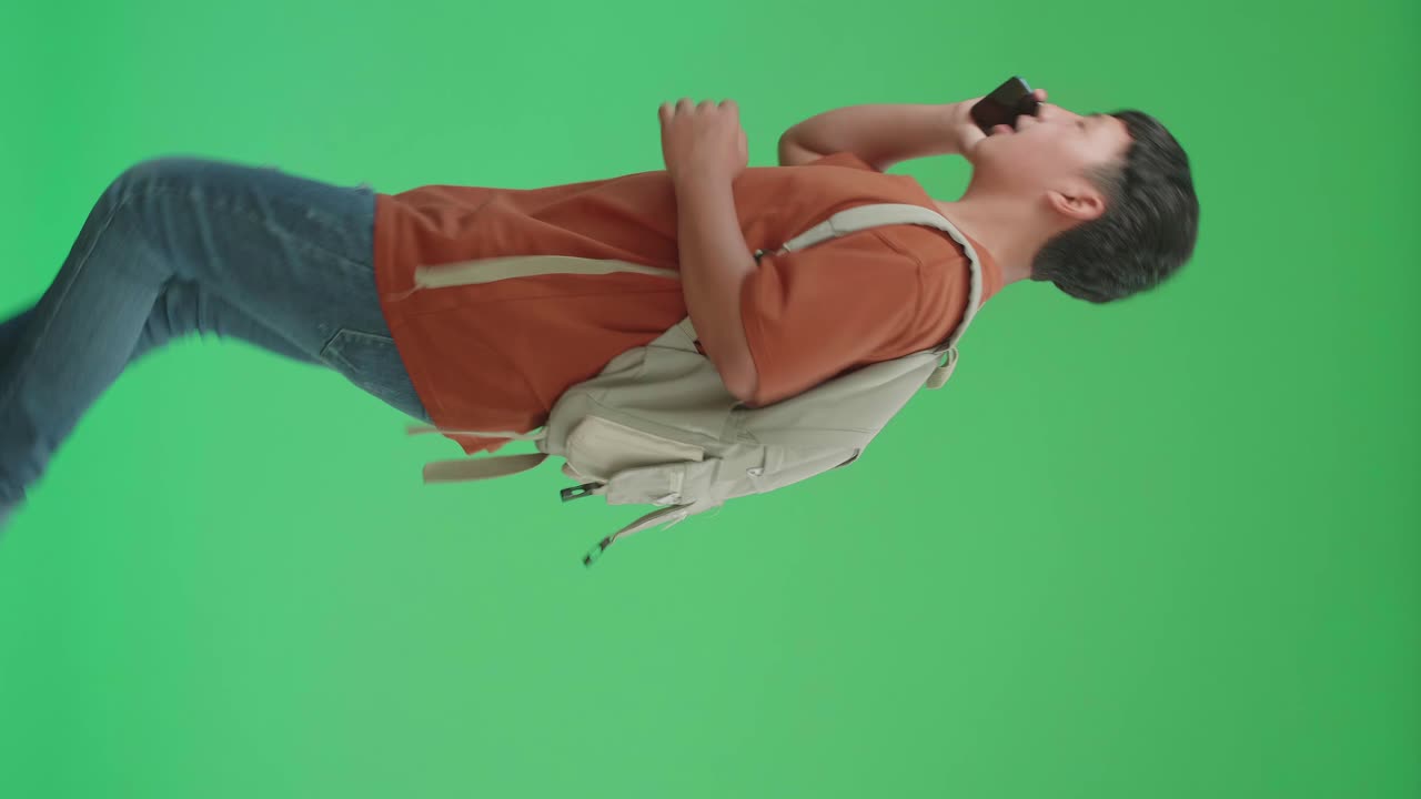 The Side View Of Asian Boy Student Wearing A Backpack, Brown T-Shirt, And Blue Jeans Talking On Mobile Phone While Running To School On Green Screen Chroma Key.