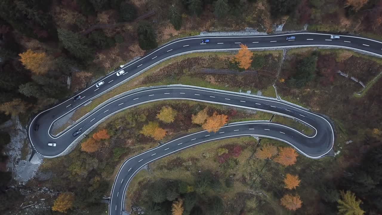 Aerial view of a winding mountain road with several sharp curves, surrounded by autumn-colored trees and forested landscape, showcasing a scenic and picturesque route through the natural terrain