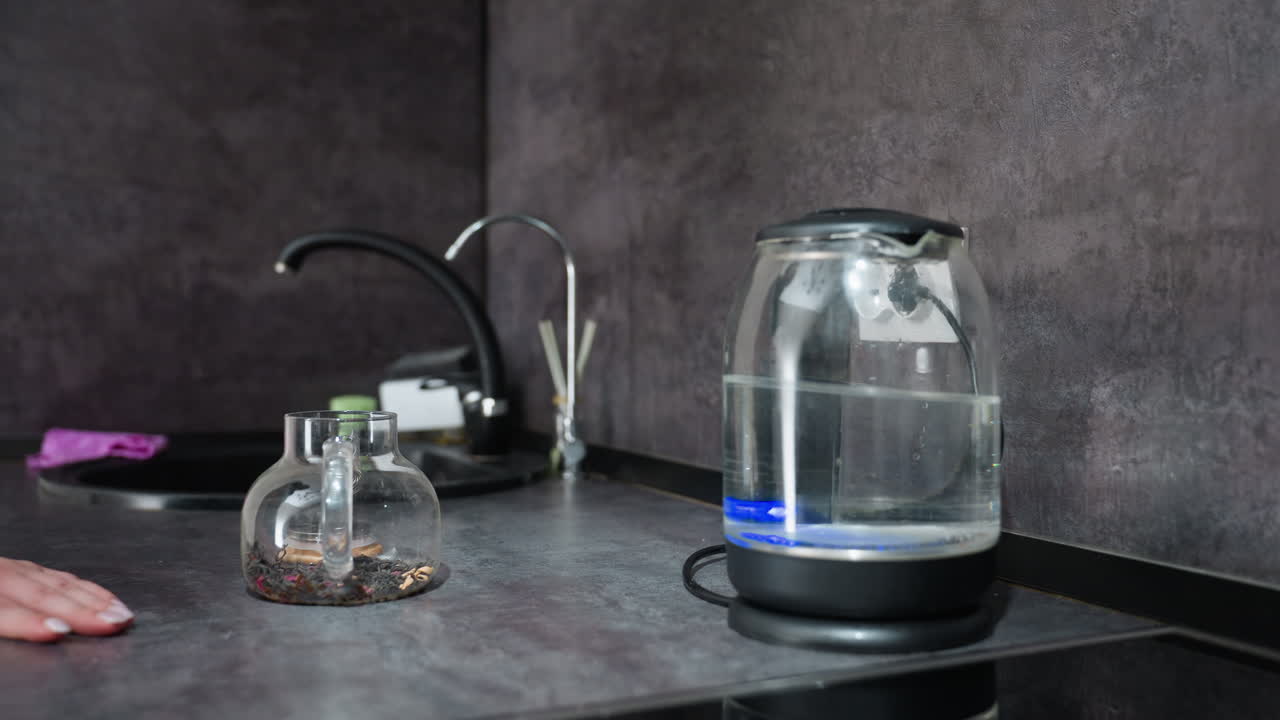 Glass Electric Kettle Heating On Countertop, Hand Nearby And Blue LED Indicator Glowing, Induction Base With Cord, Small Glass Teapot With Dried Herbs, Grey Textured Backsplash, Minimal Modern