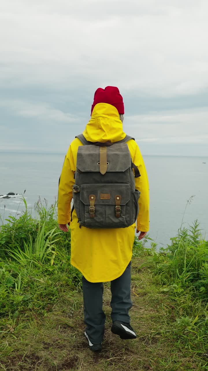 Donna escursionista con impermeabile giallo che cammina verso una vista panoramica sull'oceano