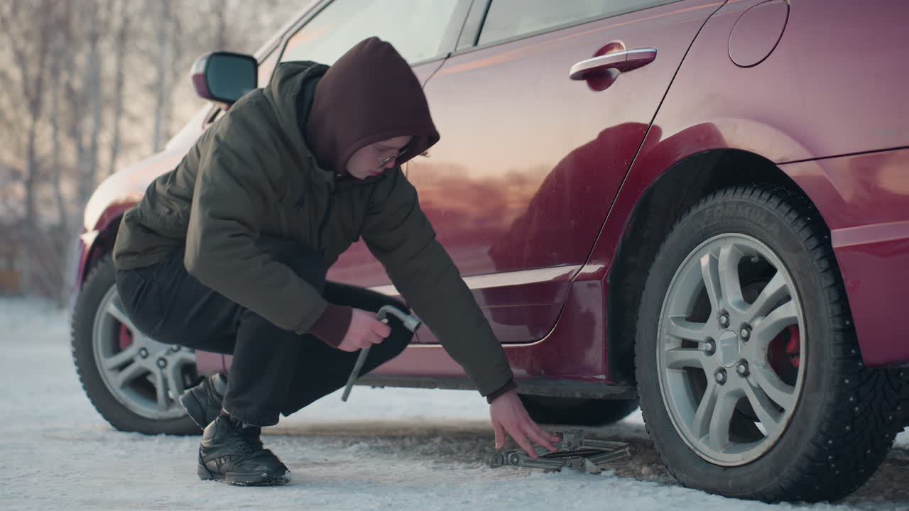 Man squats beside red car on snowy ground, positioning jack under chassis while holding lug wrench to loosen bolt on front wheel, winter scene with cold weather gear and soft daylight reflections