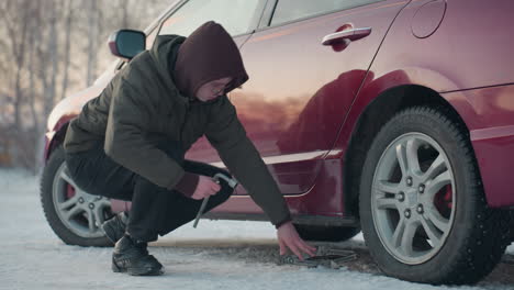 man squats beside red car on snowy ground, positioning jack under chassis while holding lug wrench to loosen bolt on front wheel, winter scene with cold weather gear and soft daylight reflections
