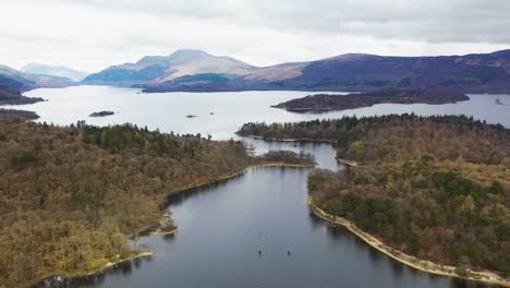 ángulo-Aéreo-De-Drones-En-Cámara-Lenta-De-Barcos-Que-Viajan-A-Través-De-Los-Estrechos-En-Loch-Lomond,-Escocia,-Con-Luss-Y-Ben-Lomond-En-El-Fondo-A-Principios-De-La-Primavera