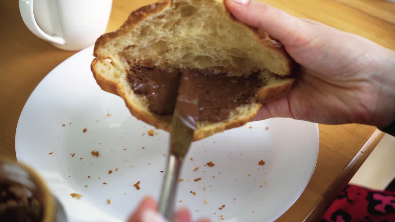 Close-up shot of woman's hands putting chocolate cream into the croissant for breakfast