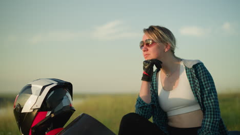 a young woman wearing a checkered shirt and crop top sits by a motorcycle, with one hand thoughtfully resting on her jaw as she gazes into the distance, her helmet rests on the bike next to her