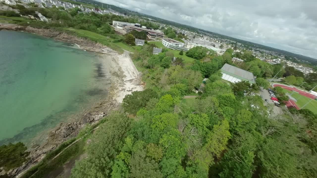 FPV drone flies above lush green trees, moving close to shoreline and large rocks under cloudy sky in Concarneau, Brittany, France, rugged coastal beauty.