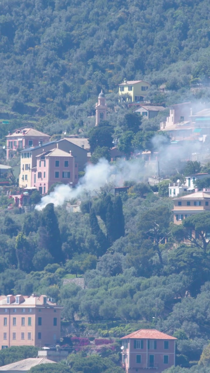 Fumo che sale dalle case in un villaggio su una collina verde.