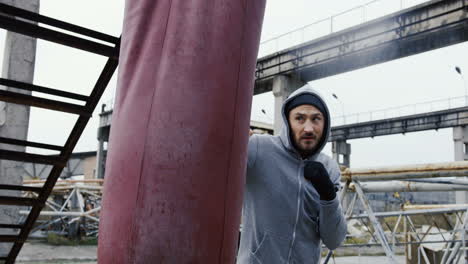 side view of caucasian bearded caucasian man in grey hoodie hitting a punching bag outdoors an abandoned factory on a cloudy morning