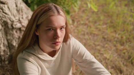 young adult seated on ground under tree, hand resting on knee, looking up contemplatively, dry foliage and earthy tones in background