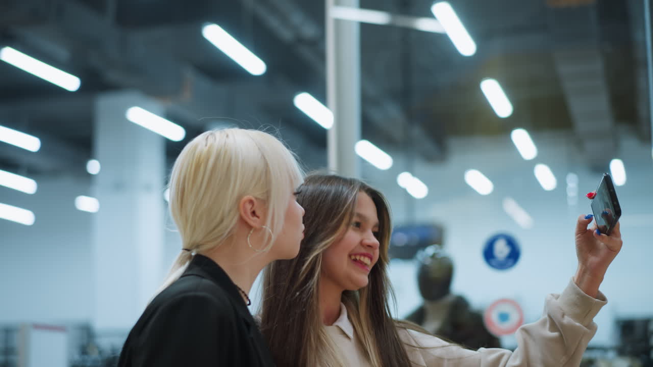 Side view of two girls smiling while taking selfie inside modern shopping mall, smartphone held forward as they pose together under bright lights with blurred retail background