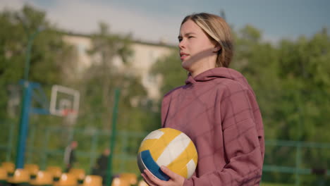 lady in maroon hoodie holding volleyball, interacting with someone in background with her hand, background features trees and building, capturing outdoor sports atmosphere and volleyball training