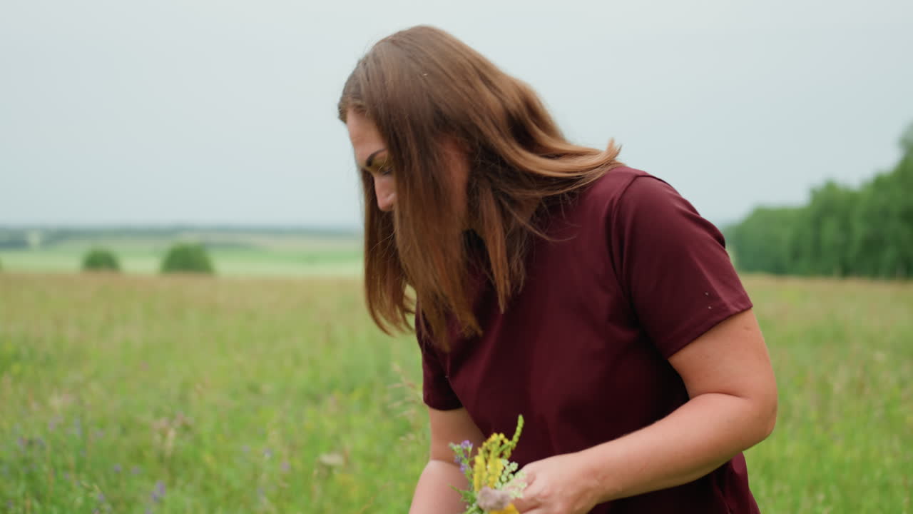 코카서스 인종 여성이 현장 학습 중 초원 식물을 채집하고 있어요. 자주색 셔츠를 입고 줄기와 꽃가루를 자세히 관찰하며 흐린 날씨에 집중한 표정으로 일지를 쓰고 샘플을 채취하는 분위기는 야외 생태 연구와 발견의 느낌을 줍니다.