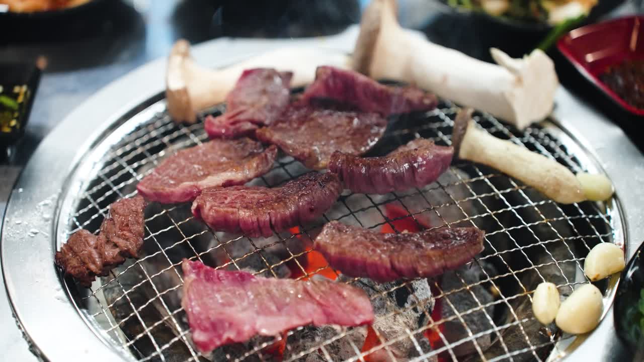 Very close-up, low-angle shot of thick cuts of Hanu beef and whole king oyster mushrooms cooking on a hot metal grate over glowing charcoal embers
