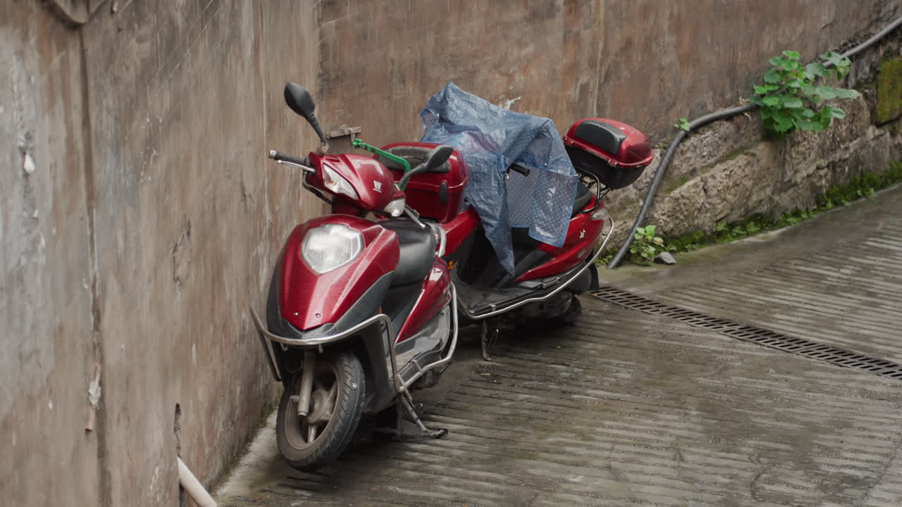 Scooter rosso incatenato in un vicolo bagnato, copertura di plastica sul sedile, pesante lucchetto a catena che assicura il veicolo contro un muro di cemento muschioso, pavimentazione in pendenza con pozzanghere e sporcizia, l'atmosfera nuvolosa evoca solitudine
