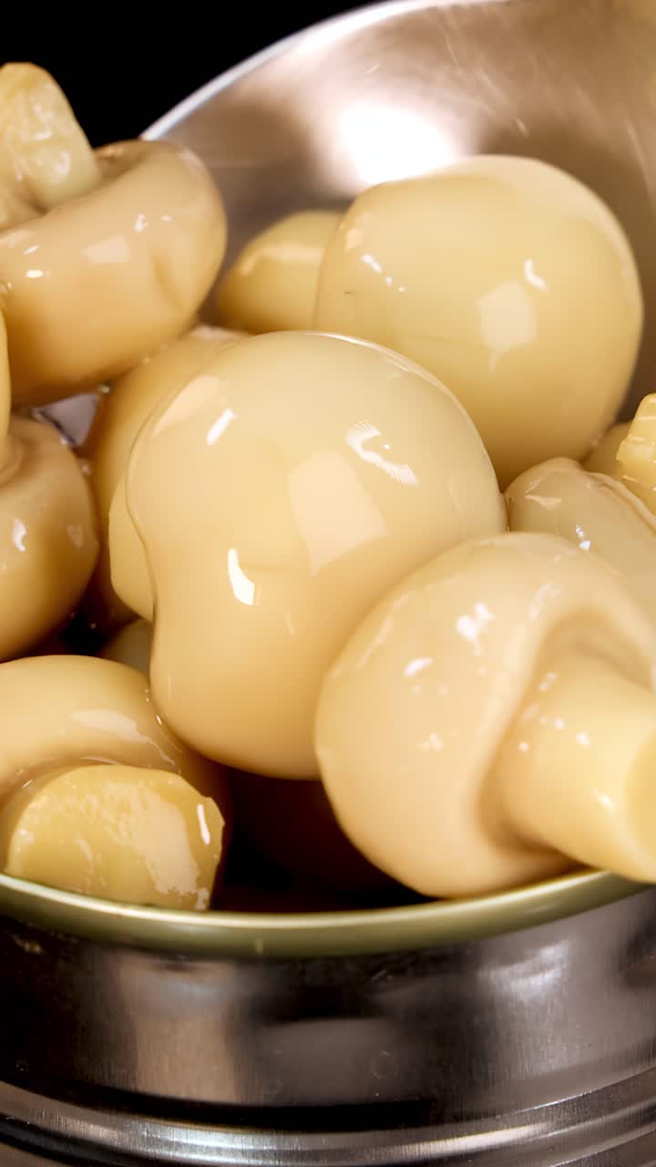 Un cucchiaio di metallo solleva funghi champignon interi e lucidi da una lattina aperta piena di salamoia, sotto una luminosa illuminazione da studio e in primo piano.