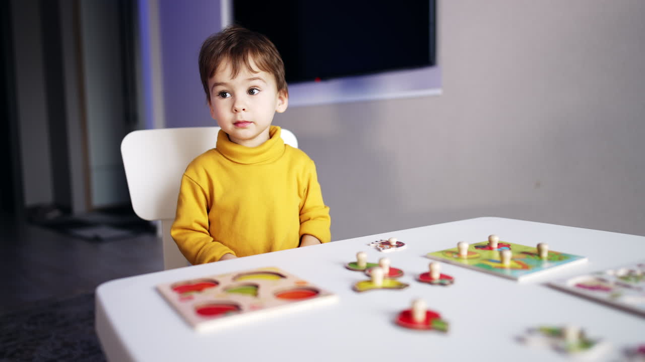 Cute smart little boy sitting at the desk. Teenage kid taking two pieces of puzzles and taps them together. Blurred backdrop.