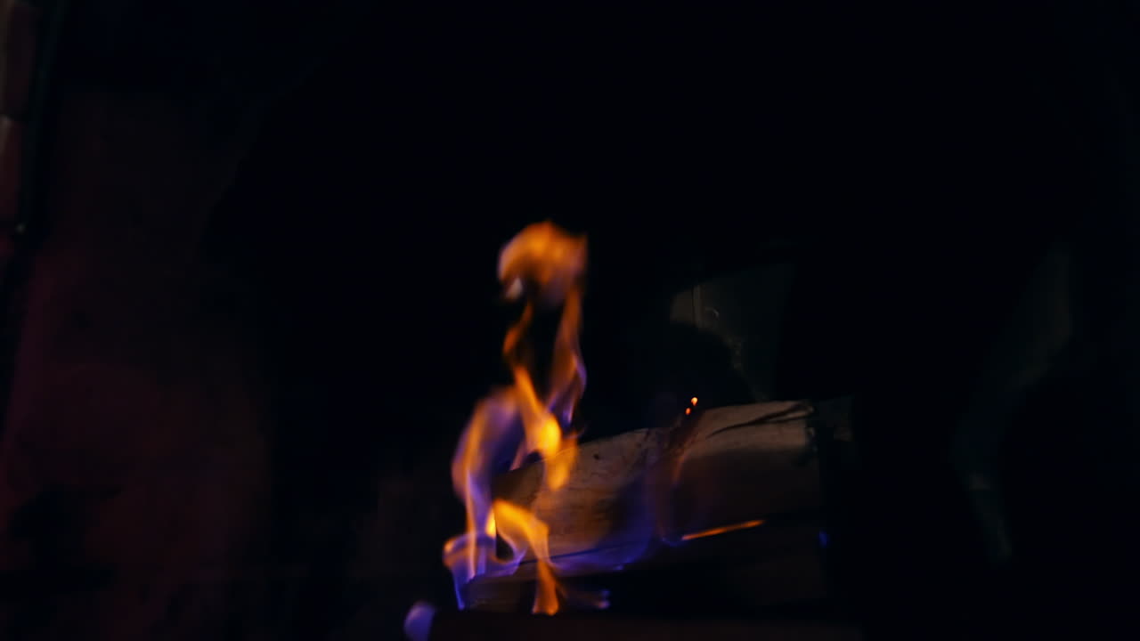 Blue and yellow fire flame burning in a stone fireplace. Wooden logs are burning in darkness. Cozy and warm atmosphere.