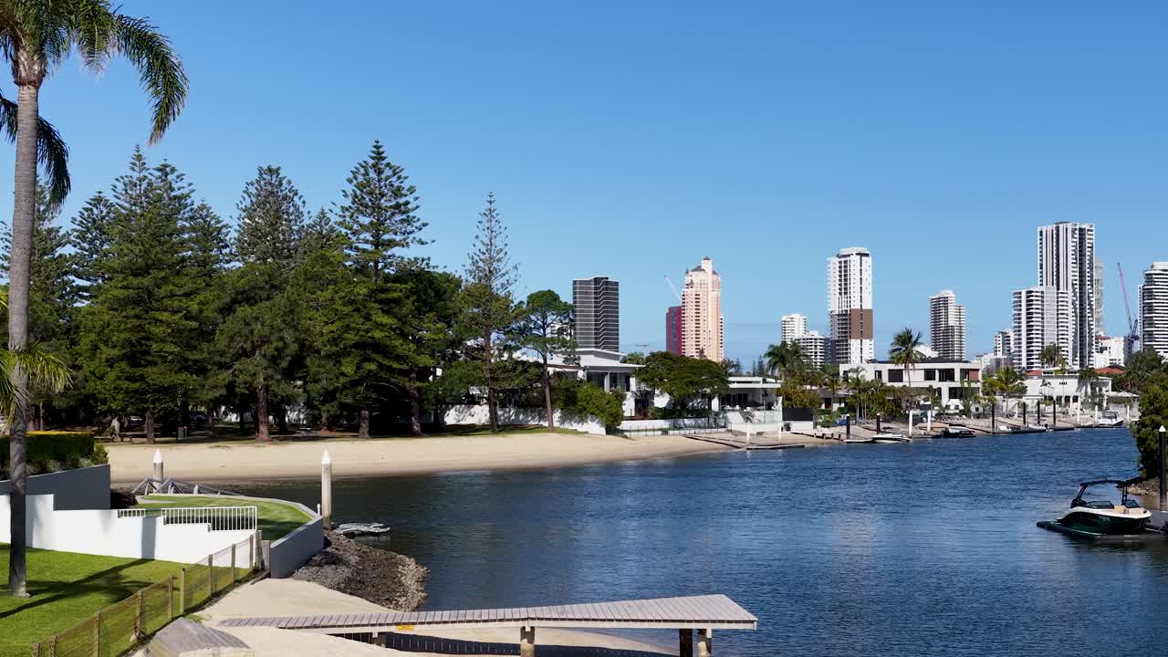 Daytime camera pan reveals canal, modern houses, palm trees, city skyline, and clear blue sky