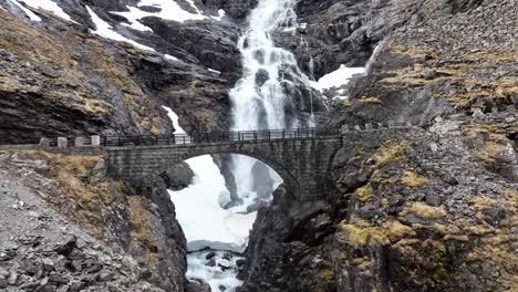 Stigfossen-waterfall-bridge-along-Trollstigen-road-fv-63-in-Rauma-Romsdal-Norway---Rising-aerial-showing-beautiful-old-stone-bridge-with-waterfall-and-snow-patches-in-background