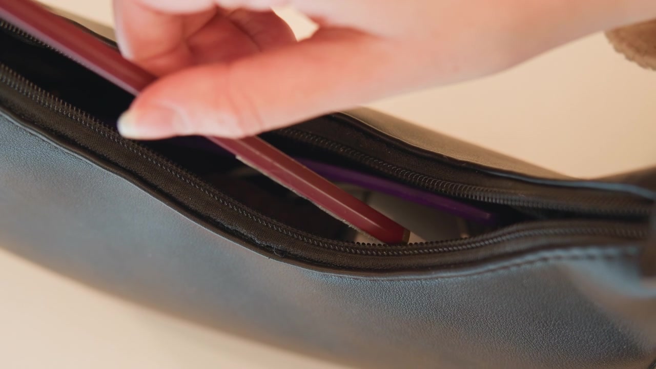 Primer plano que muestra una mano guiando un lápiz delgado dentro de una bolsa de cuero texturizado parcialmente abierta, con el deslizador de la cremallera desplazado a un lado, los dientes metálicos expuestos y el forro morado visible en el interior.