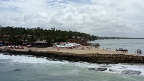 Dolly-in-aerial-drone-shot-of-the-tropical-Tibau-do-Sul-beach-in-high-tide-with-waves-crashing-into-rocks,-tourists-swimming-and-enjoying-shade-under-colorful-umbrellas-in-Rio-Grande-do-Norte,-Brazil