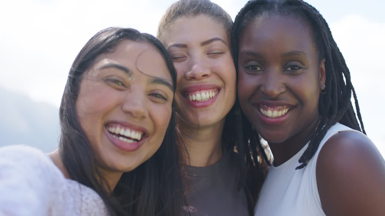 Drei afroamerikanische Frauen beugen sich zum Telefon, lächeln und richten ihre Haare für ein Selfie im Freien zurecht.