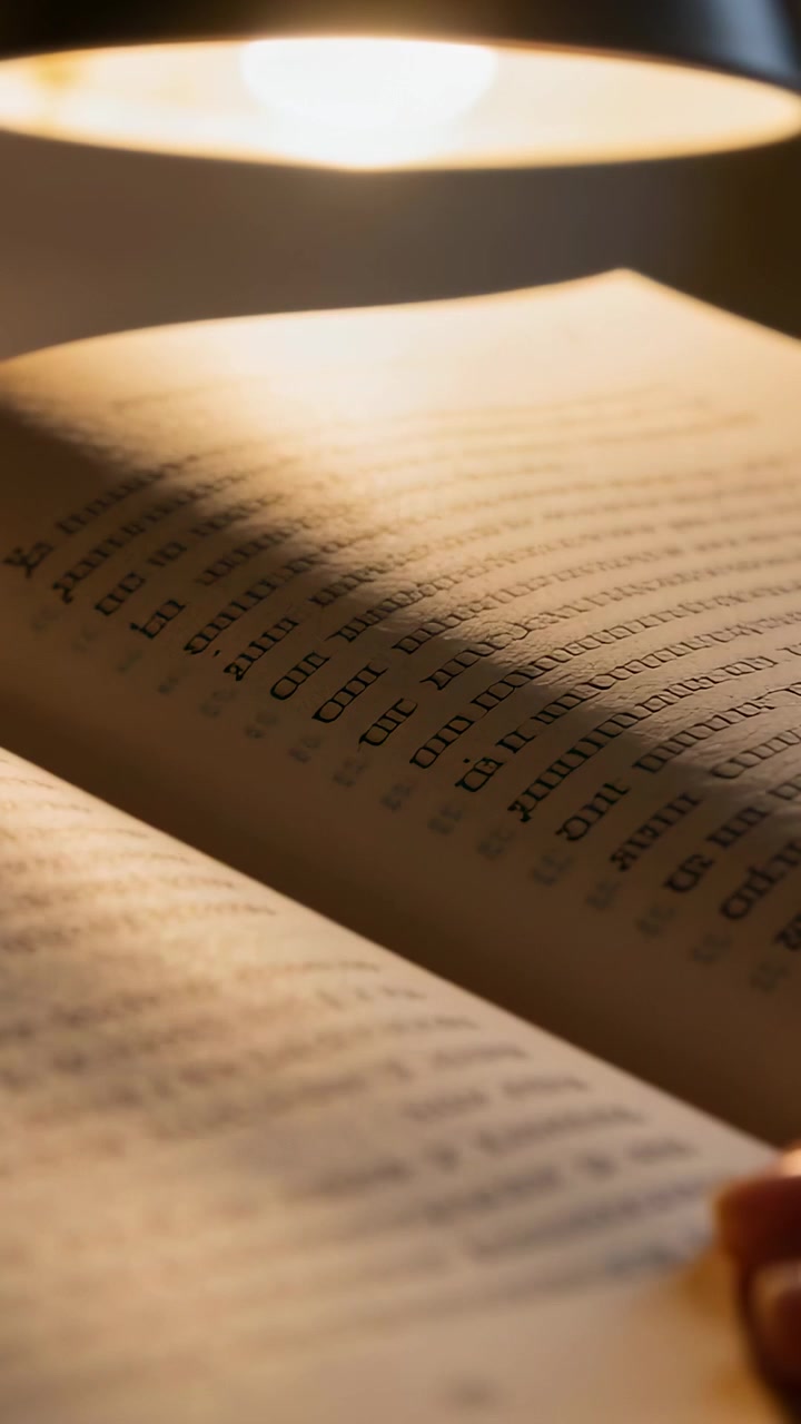 Video verticale: Dito che gira la pagina di un libro tascabile aperto per leggere a letto, sotto una lampada calda.