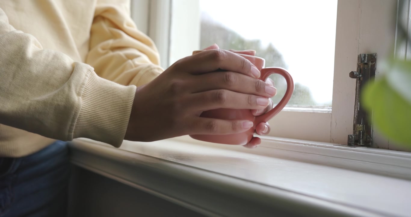 African-American woman in yellow sweatshirt cradling pink mug at sill, lifting to sip, copy space