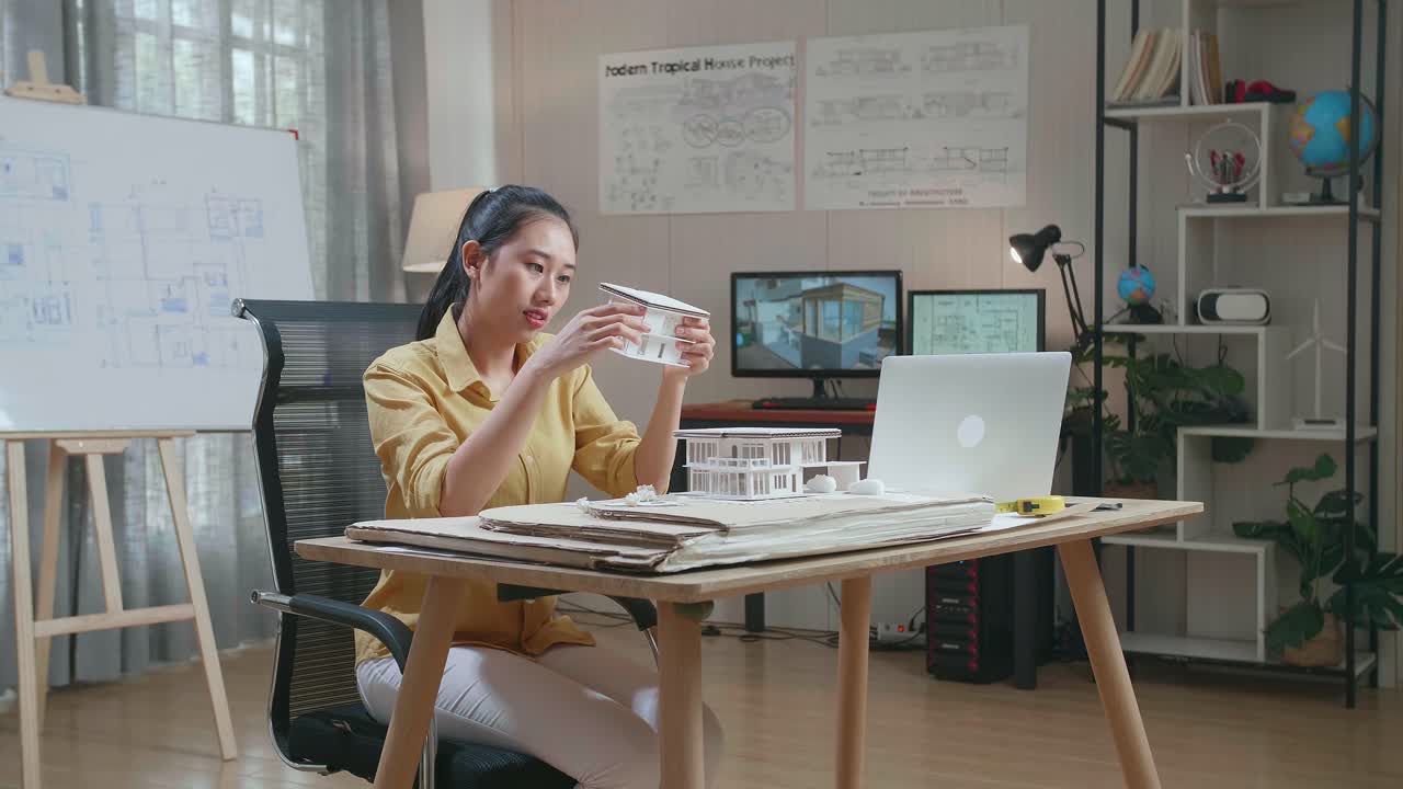 Asian Woman Engineer With A Laptop Making Paper Model Of House At The Office