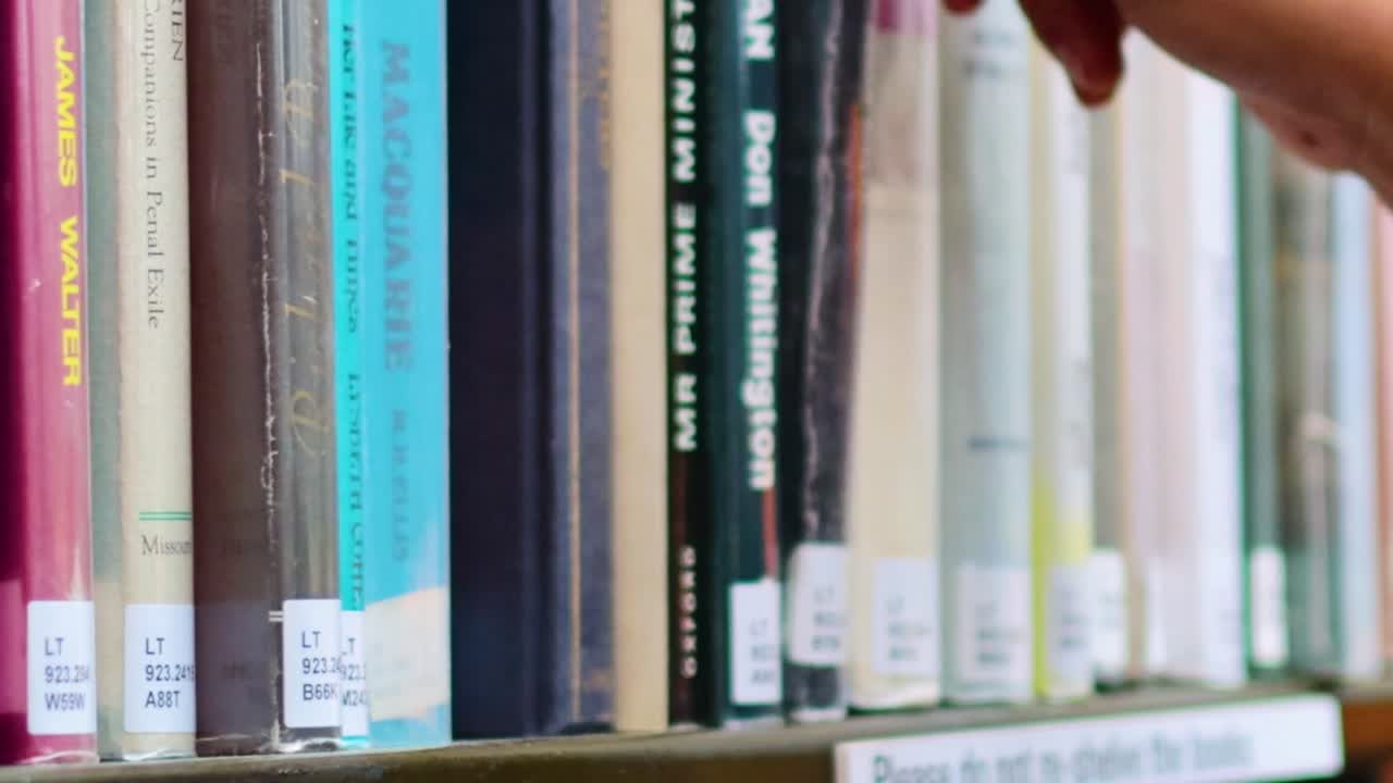 A hand reaches for a book among neatly organized library shelves, highlighting the selection process.
