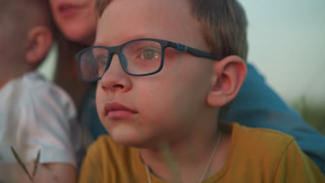 una toma de primer plano de un niño que lleva una camisa amarilla y gafas, profundamente enfocado en lo que está observando. en el fondo, una mujer y un niño más joven están suavemente borrosos