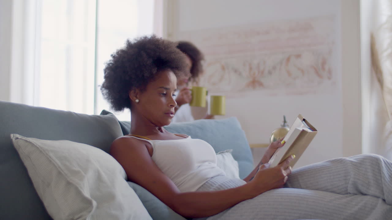 Black woman lying in bed reading when her daughter bringing tea