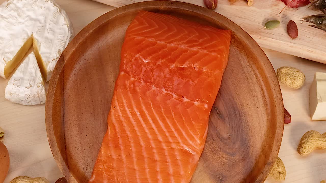 Close-up of a salmon fillet on a wooden plate, surrounded by cheese and assorted nuts.