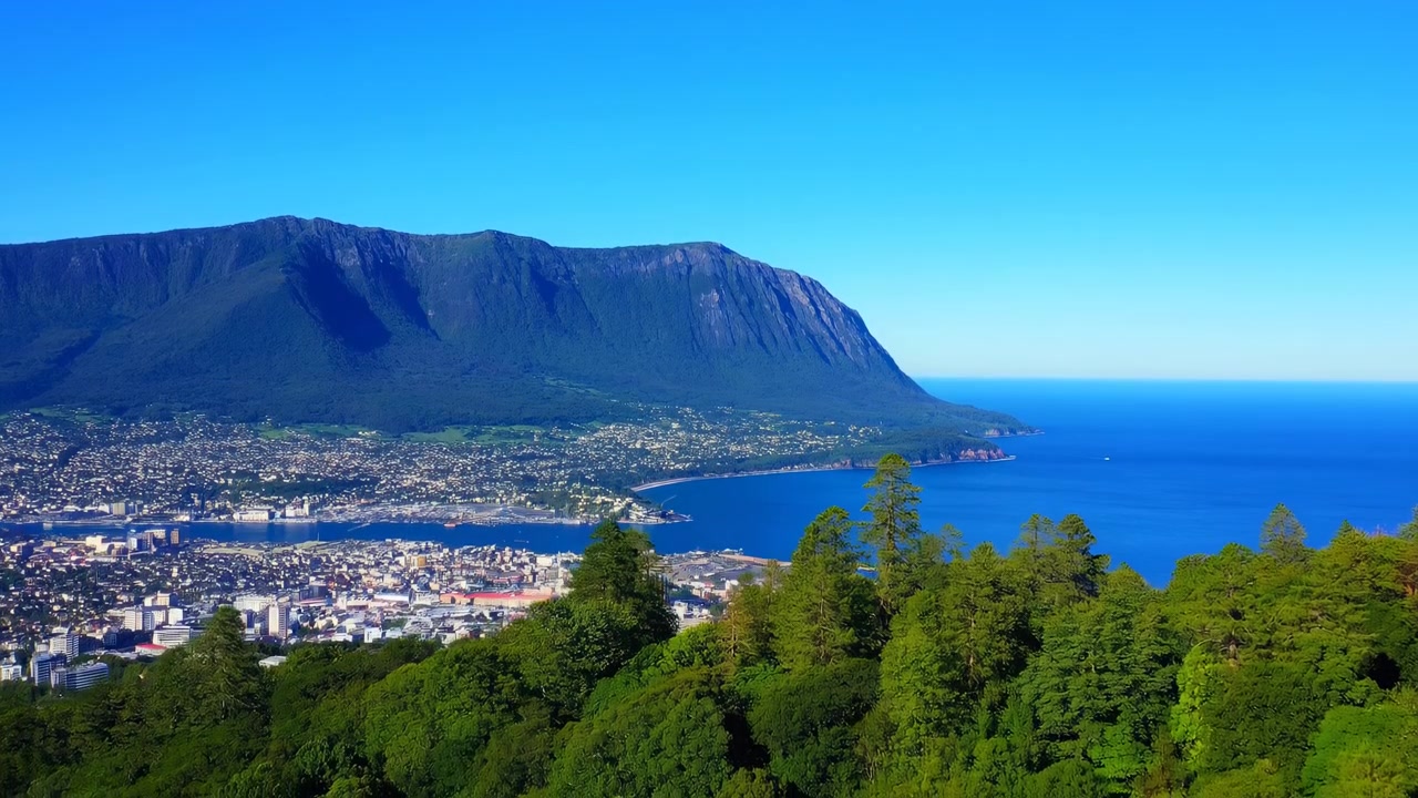 Scena in movimento dall'alto di una città costiera ai piedi di una ripida e scura catena montuosa, con una densa foresta di conifere in primo piano, un lungomare urbano compatto e una baia sabbiosa curva in secondo piano, e un'ampia