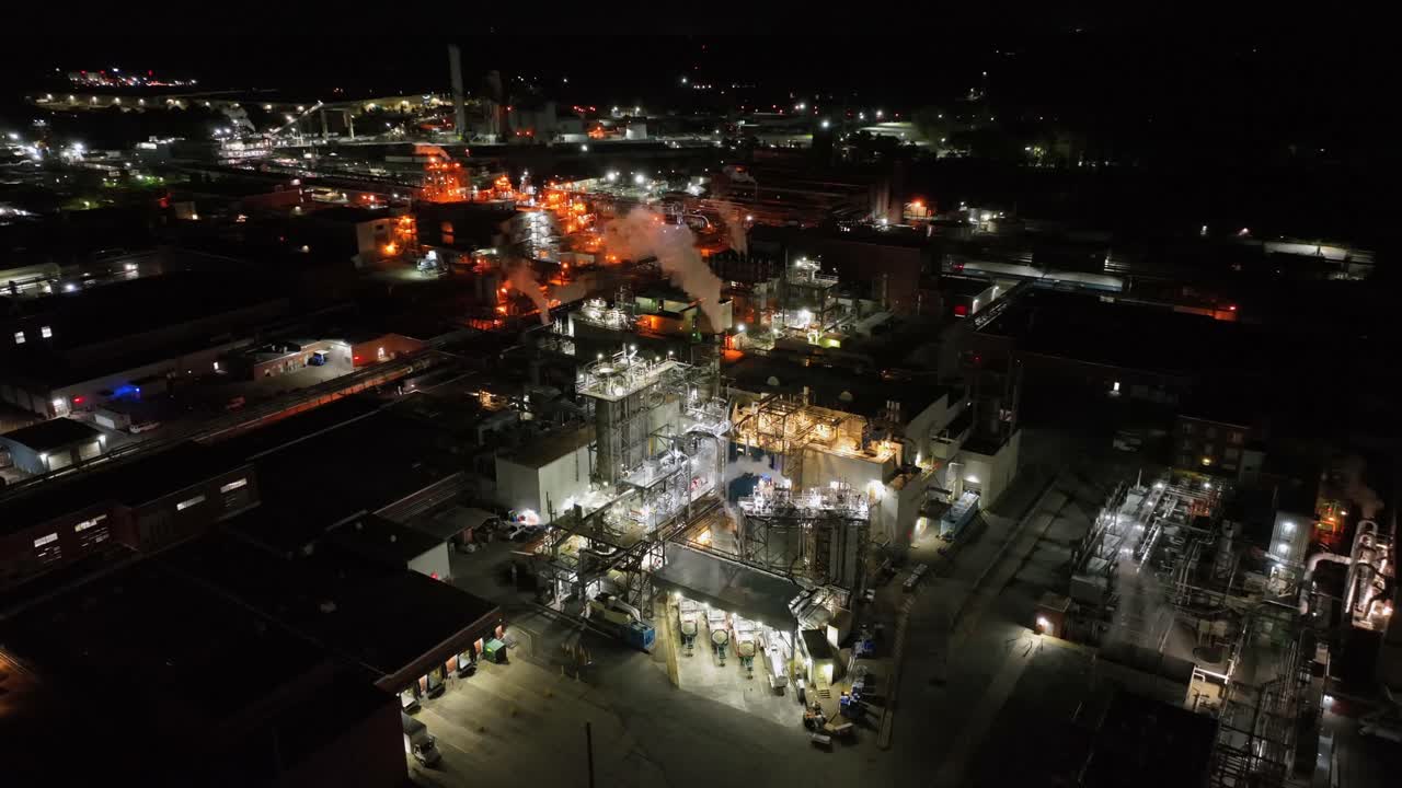Nighttime drone glide over a sprawling industrial complex with bright lights, active steam vents, and detailed machinery spread across a large energy and manufacturing facility