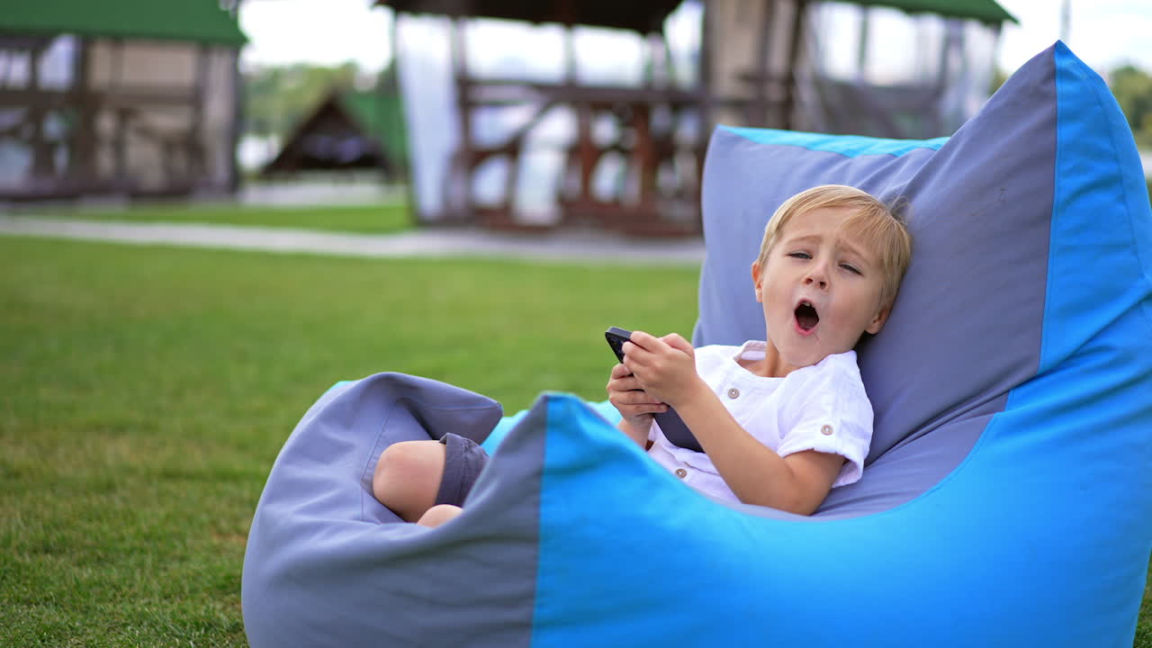 Lovely blond preschool boy sitting in bean bag chair with phone in his hands. The kid distracts for a moment saying something and turns back to gadget. Blurred backdrop.