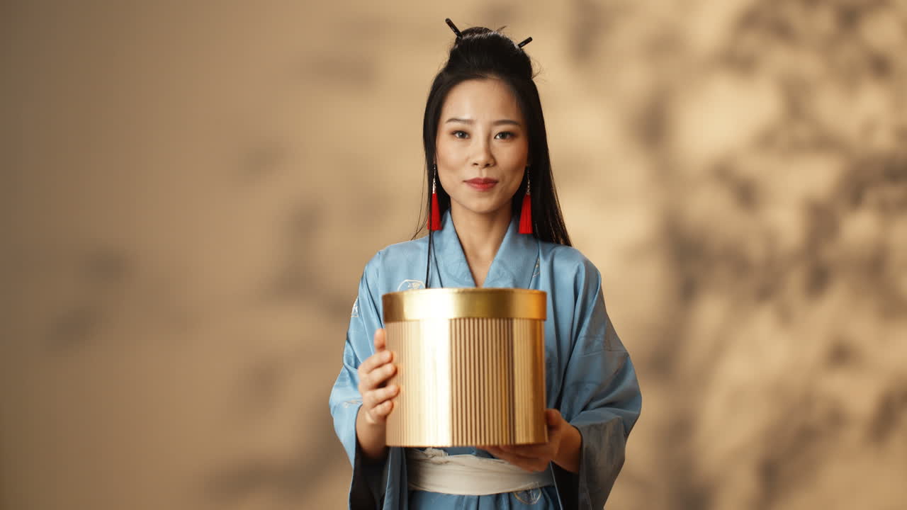 молодая азиатская женщина в голубом кимоно держит золотую коробку и показывает ее на камеру