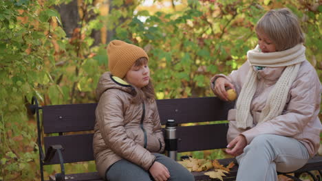 mother hands apple to daughter sitting quietly on park bench in autumn forest, daughter looks at mother with curiosity, atmosphere warms as she receives fruit with soft smile under golden leaves