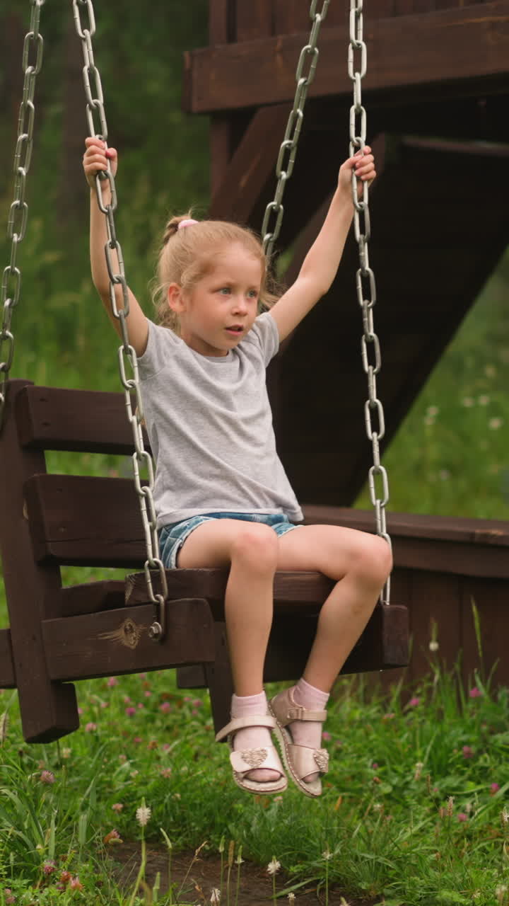 Cute little kid with braid plays wooden swings with chains on hotel playground on fresh lawn on dull summer day at countryside slow motion
