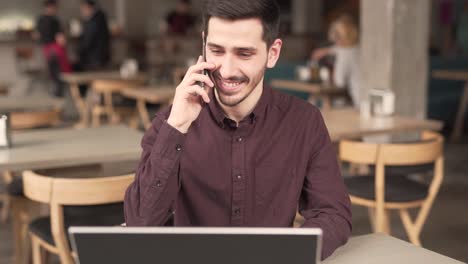 un joven apuesto con barba y bigotes está sentado en un café, bebiendo un café con leche, hablando por teléfono y sonriendo