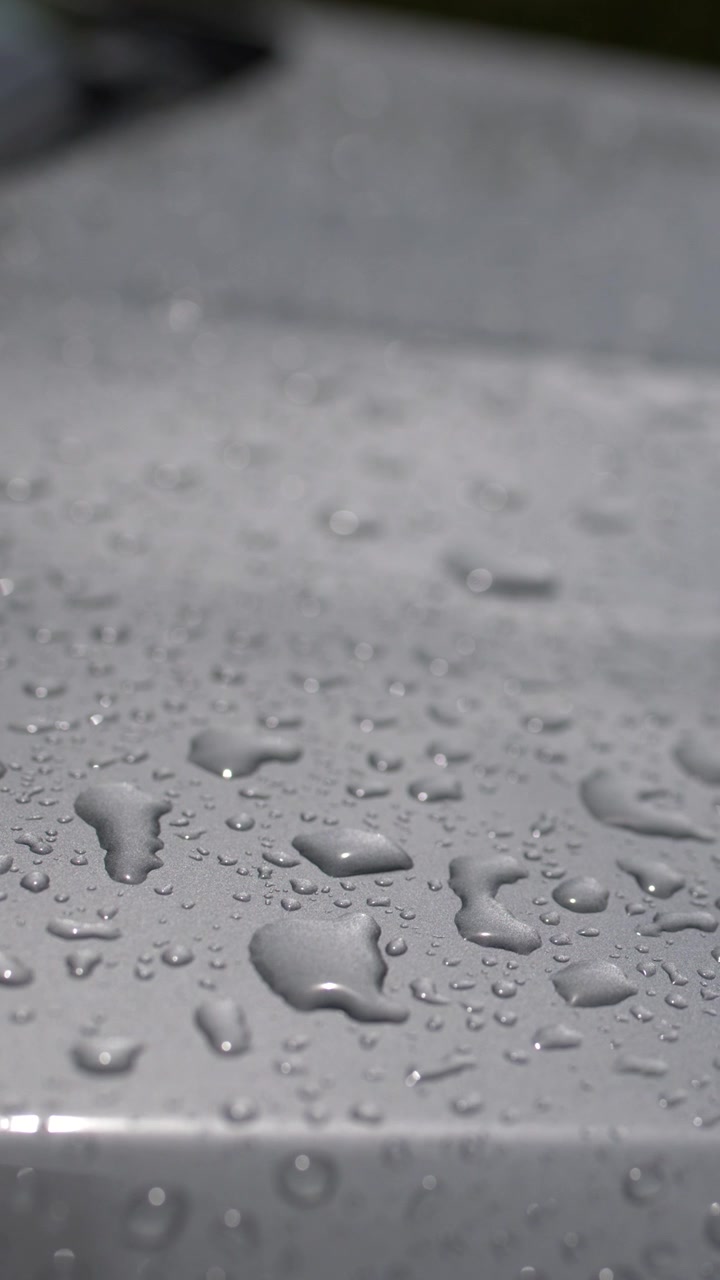 Close-up macro shot of glistening raindrops and water beads on a silver car hood.