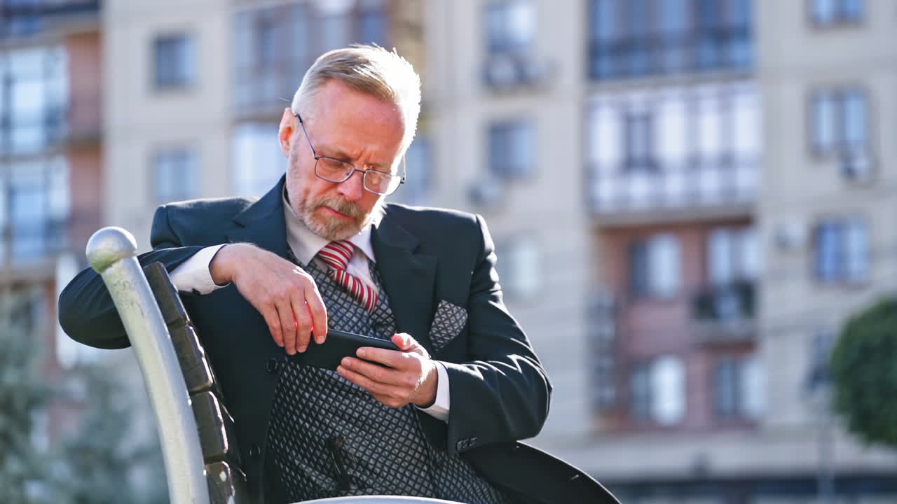 Mature businessman with beard and glasses sitting on bench in the city. Middle aged man looking seriously into his mobile phone outdoors.