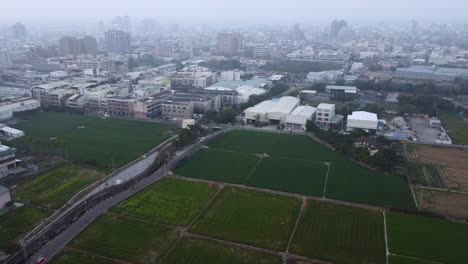 Campos-De-Arroz-Verdes-Junto-A-Una-Zona-Urbana-Bajo-Un-Cielo-Brumoso,-Un-Toque-De-Agricultura-Urbana,-Vista-Aérea