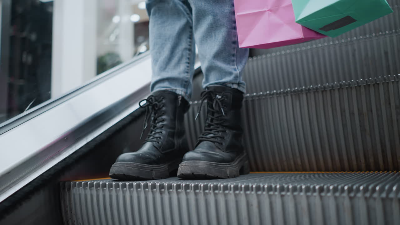 close-up de pernas em botas pretas e jeans de pé perto do corrimão da escada rolante enquanto segura sacos de compras em um centro comercial vibrante, a cena captura uma experiência de compras urbana elegante com detalhes modernos