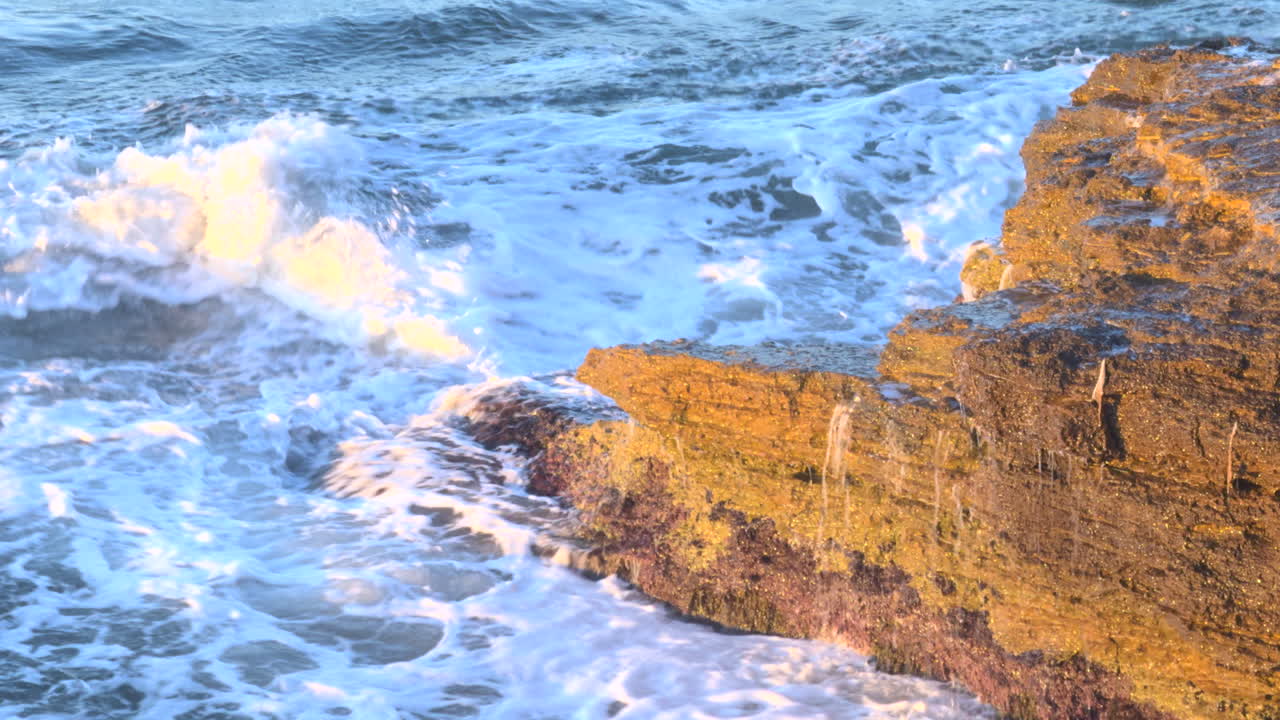 Close-up of a crashing wave hitting a rocky coast at sunset. Water splashes with dramatic force and golden light reflections in a breathtaking coastal seascape.