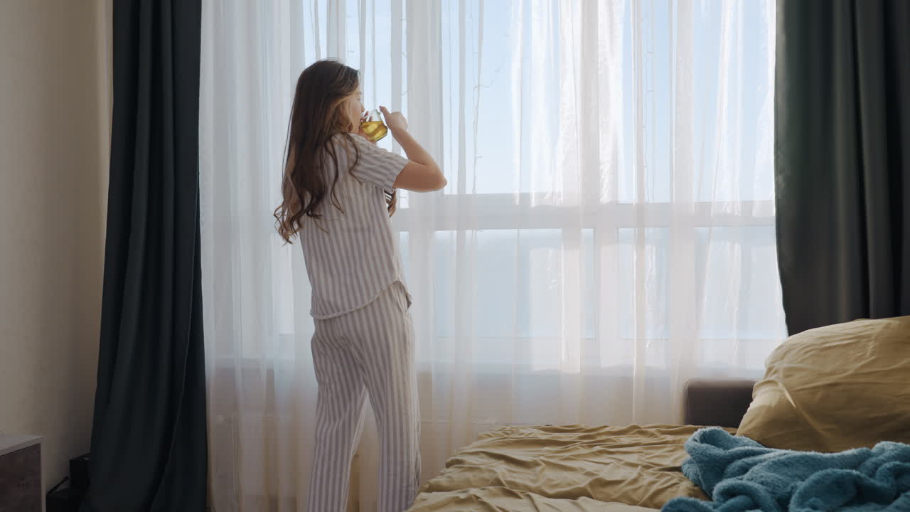 Donna che sorseggia un caffè vicino alla finestra nella tenue luce del mattino, vestiti a righe e tazza in mano mentre guarda il panorama della città, il vapore che sale e la postura rilassata suggeriscono che sia una scrittrice freelance che pianifica la giornata, delicata