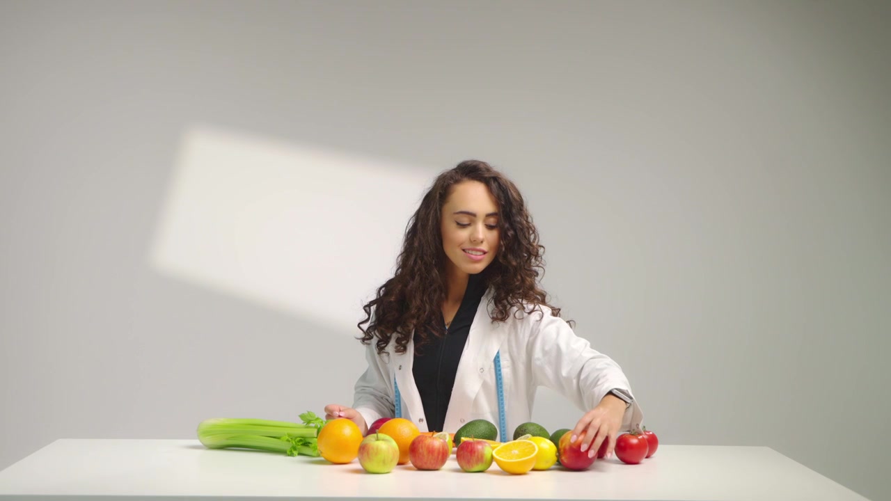 Nutrizionista donna sorridente in camice bianco esamina frutta e verdura fresca su una scrivania su sfondo bianco. Concetto di alimentazione sana, pianificazione della dieta e benessere al rallentatore.