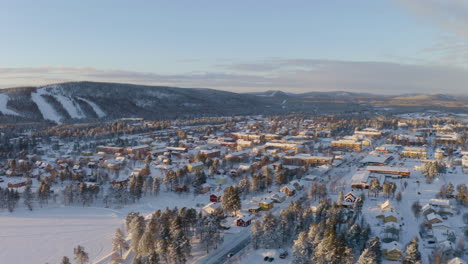 Luftbild-über-Die-Riesige-Skandinavische-Schneebedeckte-Siedlung,-Umgeben-Von-Bewaldeten-Berglandschaften-Unter-Sonnenaufgangs-Skylinen