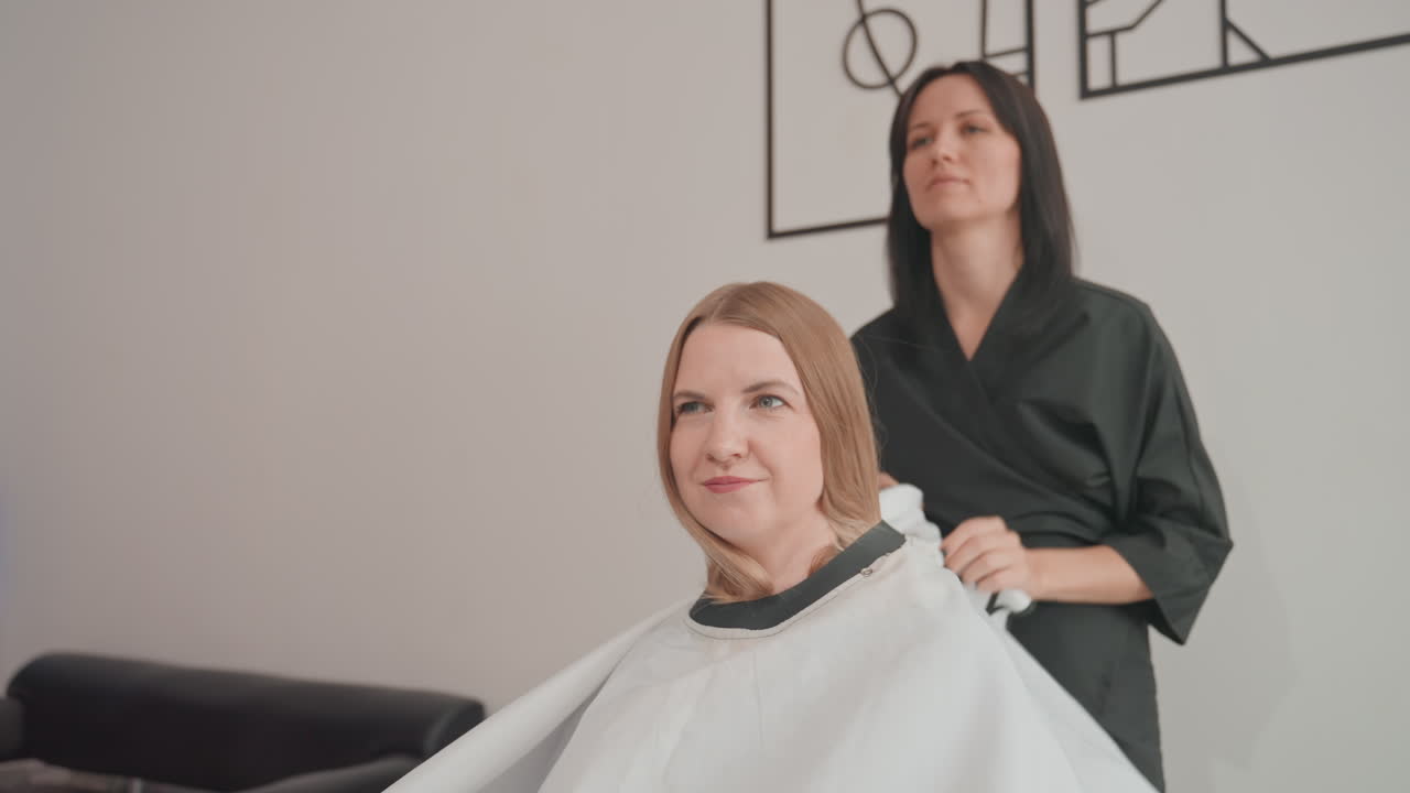 Donna bianca bionda che riceve un taglio di capelli in un salone, stilista che drappeggia una mantellina protettiva e liscia i capelli di media lunghezza, pettinandoli e dividendoli in sezioni per un taglio preciso, cliente sorridente e rilassata in un ambiente moderno e neutro